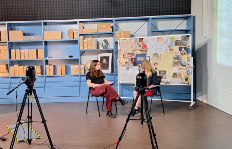Two people sit in a broadcast studio in front of a board featuring cut out records and images. They are facing two cameras.